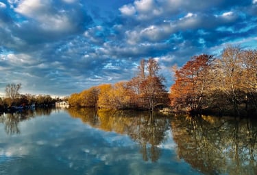 Tranquil river landscape with autumn trees reflecting in calm water under a dramatic blue cloudy sky.