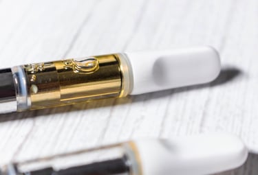 Close-up of a glass cannabis oil vape cartridge with gold distillate and white mouthpiece on a light wood surface.