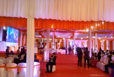 Indoor wedding reception with orange and white decorations, guests seated on white chairs.
