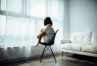 Eine jugendliche Person sitzt zusammengekauert auf einem Stuhl und blickt aus dem Fenster.