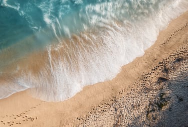 Aerial view of foamy turquoise waves crashing onto sandy beach with footprints along the shore.