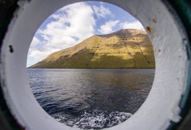 View of a green mountain and blue ocean through a circular porthole window on a boat.