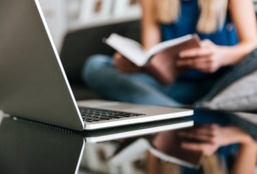 A silver laptop sits on a table while a woman studies a book in the background.