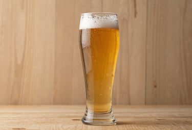 A high-end studio shot of a tall beer glass filled with a golden ale, condensation on the glass. The background is a textured Earthy Tan wood, softly lit.