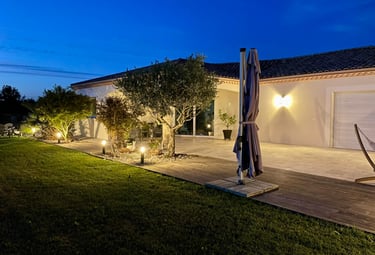 Modern luxury villa patio at night with warm outdoor lighting, garden trees, and a wooden deck.