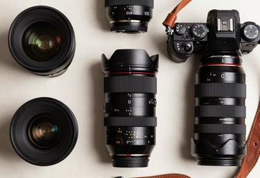 A high-contrast flat lay of professional photography equipment including lenses, a jet black camera body, and leather straps on an off-white background. Global / Western studio environment.