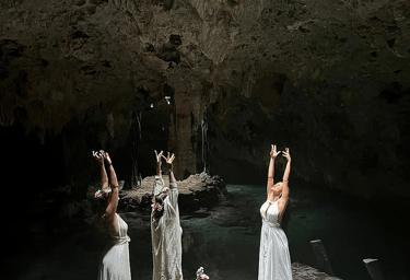 Three women in white dresses performing a sacred womb dance ritual in a cenote cave in Tulum, Mexico