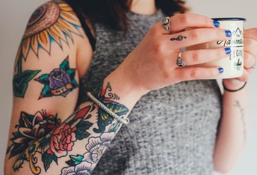 A woman with multiple tattoos, multiple rings and blue fingernails holds a coffee mug