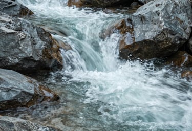 An artistic close-up of a mountain stream flowing over smooth rocks in a North American forest. The lighting is soft and diffused. The image emphasizes a clean and modern aesthetic with natural textures in teal grey and mist white.