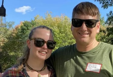 A smiling couple wearing sunglasses posing outdoors on a sunny day with autumn trees in the background.