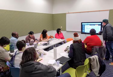 A group of people gathered around a table, looking intently at a presenter in the front of the room