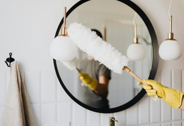 Cleaning a mirror with care — a gloved hand holding a duster in a bright, clean bathroom