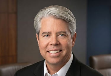 Professional headshot of a smiling male executive with grey hair in a black suit jacket and white shirt.