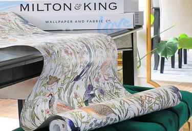 Milton and King floral bird wallpaper roll draped over a green velvet ottoman in a bright room.
