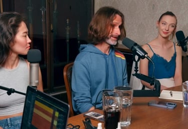 a man and woman sitting at a table with microphones doing a podcast