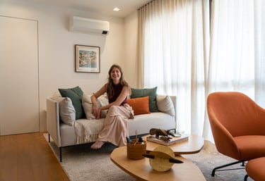 Mulher jovem sorrindo, sentada descalça em um sofá com almofadas coloridas, em uma sala aconchegante