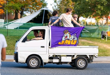 Luke Gilmore riding on the back of a Kei truck with a JMU banner on it.
