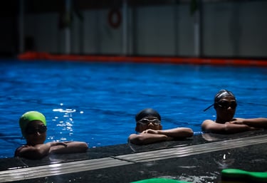 “Children wearing swim caps and goggles resting at the edge of a swimming pool.”
