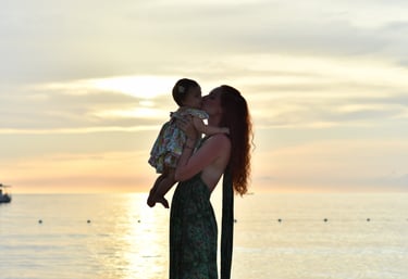 A mother holds her baby up and kisses her cheek, silhouetted by a yellow sunset.