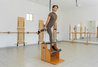 Woman performing a standing leg exercise on a wooden Pilates Wunda Chair in a studio setting.