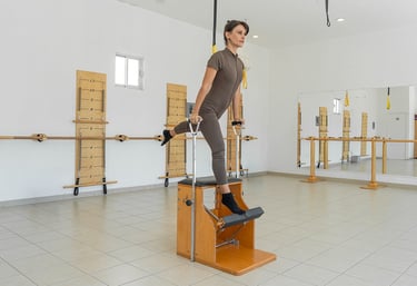 Woman performing a standing leg exercise on a wooden Pilates Wunda Chair in a studio setting.