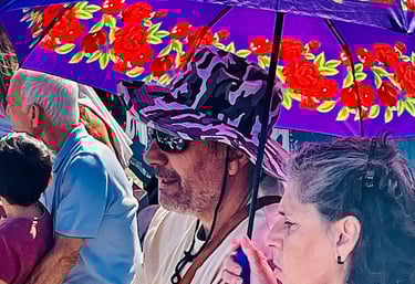 A man in a purple hat and a woman sit under a floral purple sun umbrella at an outdoor event.