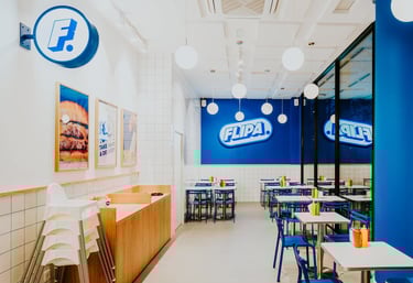 Vista de l’interior de Flipa Burger amb logotip a la paret blava, decoració retro i ambient lluminós
