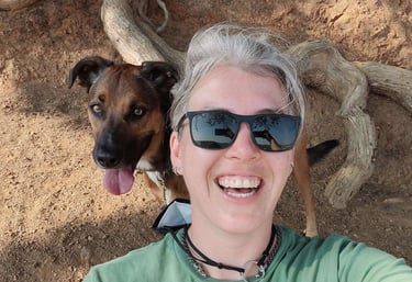 A smiling person in sunglasses takes a selfie under a tree with a dog seated smiling behind them