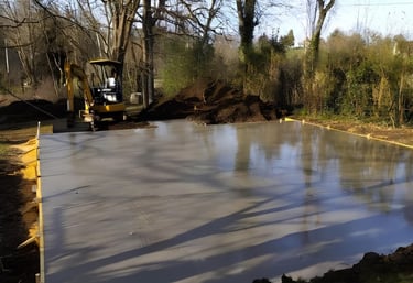 A freshly poured wet concrete slab foundation at a construction site with an excavator in the background.
