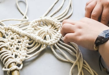 Manos elaborando un colgante de pared de macramé con cordón de algodón blanco sobre una madera.