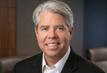 Professional headshot of a smiling male executive with grey hair in a black suit jacket and white shirt.