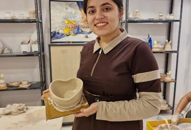 A smiling young woman holding her handmade clay bowl in a pottery classes studio with shelving.