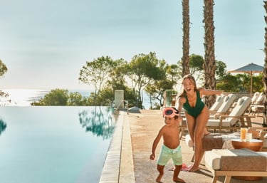 A mother and child playing by a luxury infinity pool at Ikos hotel with trees and ocean views.