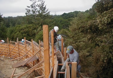 Construction workers pour concrete into a curved wooden retaining wall frame on a hillside.