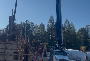 A white concrete boom pump truck and mixer pouring cement into wooden wall forms on a construction site.