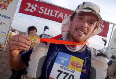 Smiling ultramarathon runner holding a gold medal at the Marathon des Sables finish line banner.
