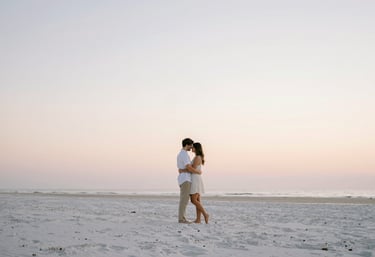 A serene photograph of a couple embracing on a quiet beach in Jacksonville at sunrise. The composition is minimalist and airy, with soft pastel colors in the sky and clean white sand. Refined and elegant style.