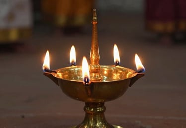 A traditional brass oil lamp with five glowing flames, casting a warm amber light in a dimly lit spiritual space, South Asian / Tamil Nadu, India.