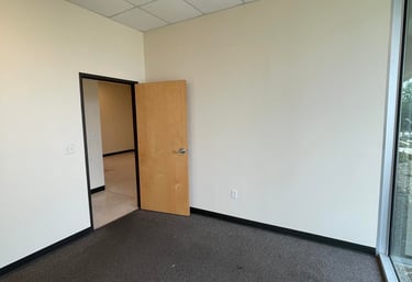 Empty commercial office space with grey carpet, beige walls, and a wooden door for rent.