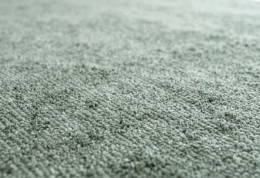 A close-up of a high-pile luxury carpet in a soft sage green (#BFCBC2), showing the rich texture and quality of the weave in soft lighting.