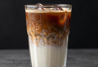 A vertical shot of a layered iced coffee in a tall glass, condensation dripping, with swirls of light beige milk mixing into dark brown espresso, modern North American / US photography style.