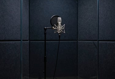 A minimalist and sophisticated vocal booth seen through soundproof glass. A professional microphone stand is silhouetted against dark navy acoustic walls. Global / International.