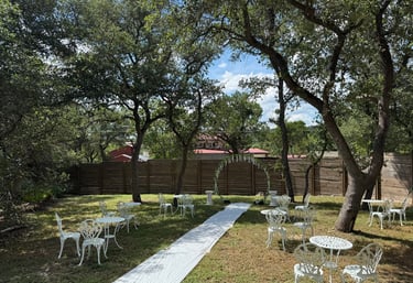 Outdoor wedding ceremony setup with white bistro chairs and an aisle runner under shaded oak trees.