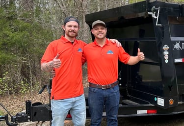Two men in orange shirts giving thumbs up by a black dump trailer for  junk removal services.