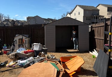 A man stands in an open backyard storage shed surrounded by cluttered outdoor items and lawn equipment.