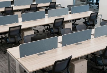 A high-angle photograph of a modern open-plan office workspace featuring rows of clean desks and ergonomic chairs in professional slate blue and white.