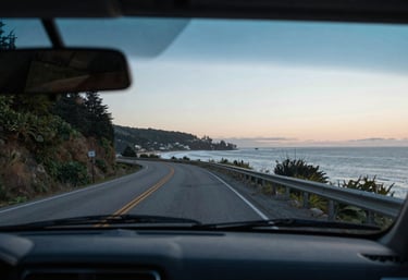 A view from inside a car through a perfectly clear, flawless windshield looking out at a scenic North American coastal road at dawn.