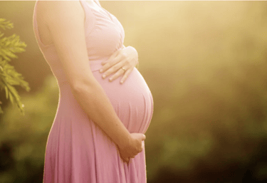 Pregnant woman in a pink maternity dress holding her baby bump outdoors in golden sunset light.