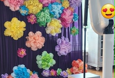Photo booth setup featuring a purple curtain backdrop adorned with colorful oversized paper flowers.