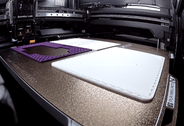 Bambu Lab 3D printer bed with purple and white filament parts being printed.